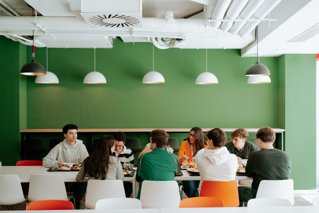 Grupo de jóvenes sentados alrededor de una mesa en una cafetería, conversando y compartiendo comida en un ambiente moderno con paredes verdes